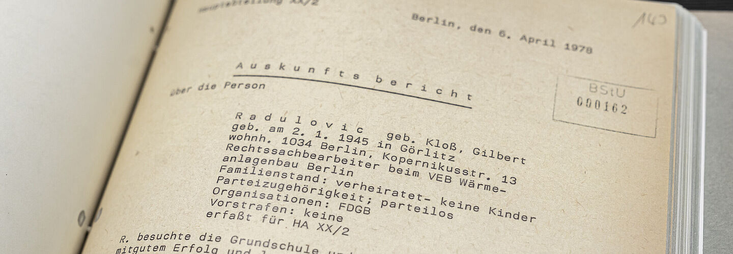 Ein Blick in die Beispielakte. Auf der aufgeschlagenen Seite sind unter der Überschrift "Auskunftsbericht" Personendaten zu Gilbert Radulovic aufgeführt. Darunter steht, dass er für die Hauptabteilung XX Abteilung 2 erfasst wurde. Am unteren Bildrand ist ein Teil eins von der Stasi verfassten Lebenslaufs über Herrn Radulovic zu sehen.